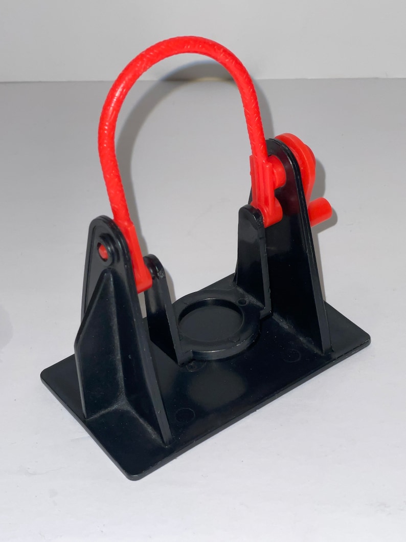 May include: A black plastic toy with a red plastic loop and a small red lever. The toy is designed to hold a small object in the centre.