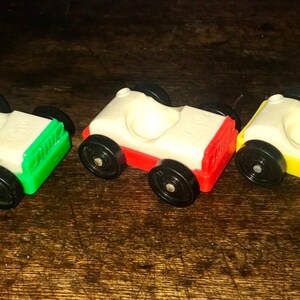 May include: Three vintage Little People toy cars. The cars are white with a green, red, and yellow bottom. Each car has a black steering wheel and four black wheels.