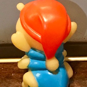Puede incluir: Una peque&ntilde;a figura de pl&aacute;stico amarilla de Winnie the Pooh con un pijama azul y un gorro de dormir rojo.
