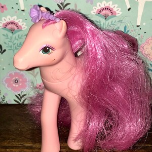 Puede incluir: Juguete My Little Pony rosa con un accesorio para el pelo de flor morada y pelo largo rosa.