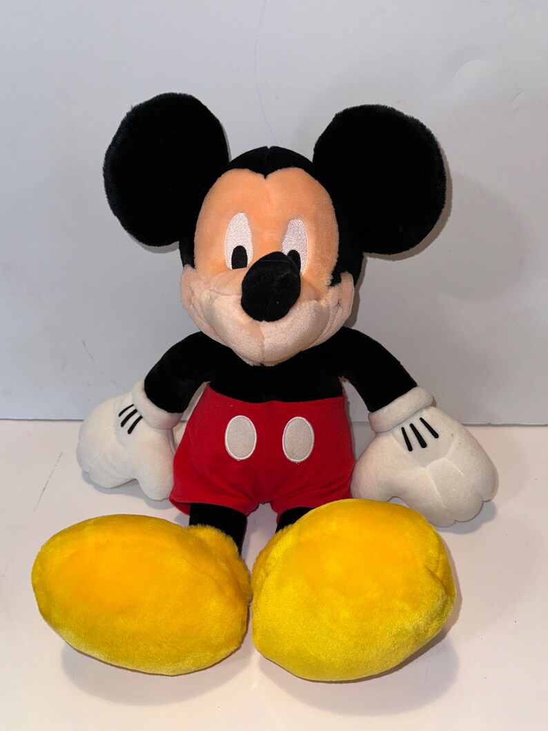 May include: A plush Mickey Mouse toy with black ears, a black nose, and red shorts with two white buttons. The toy has yellow shoes and white gloves.