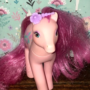 Puede incluir: Pony rosa My Little Pony con una flor morada en la cabeza y una larga melena rosa. El pony tiene ojos verdes y un dise&ntilde;o rosa y azul claro en el costado.
