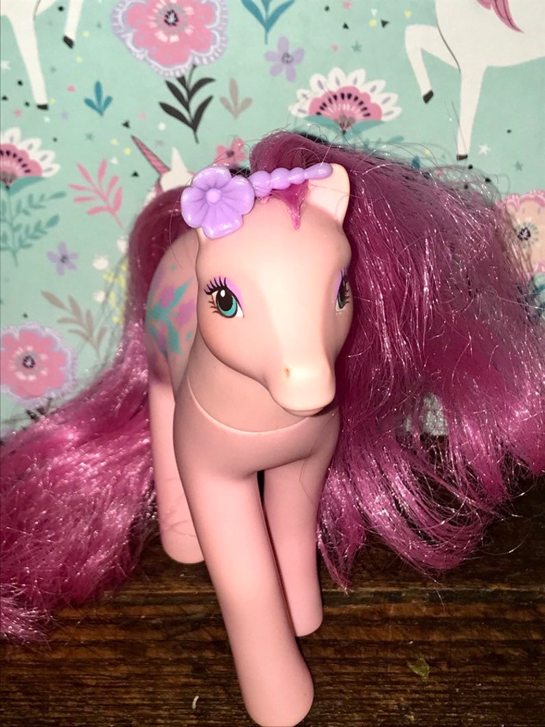 Puede incluir: Pony rosa My Little Pony con una flor morada en la cabeza y una larga melena rosa. El pony tiene ojos verdes y un dise&ntilde;o rosa y azul claro en el costado.