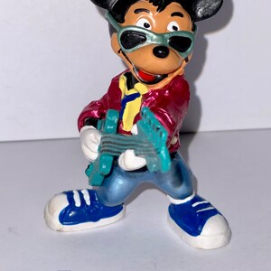 May include: A plastic figurine of Mickey Mouse wearing sunglasses and a red jacket, playing a blue electric guitar.