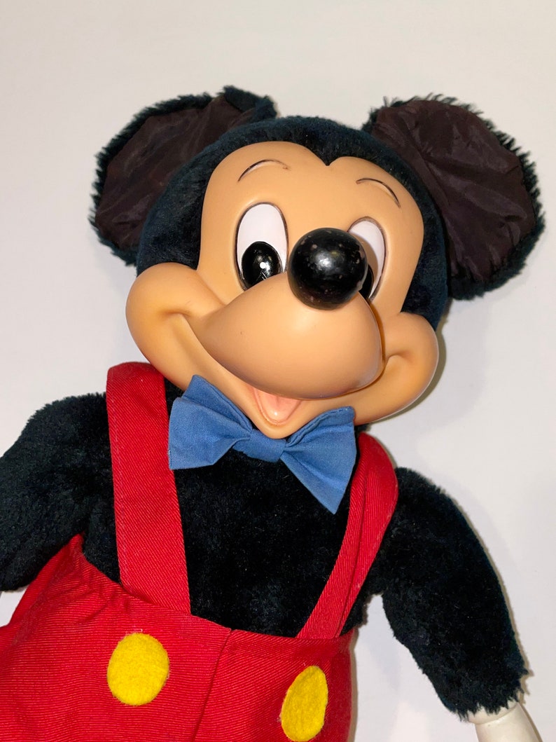 May include: A plush Mickey Mouse toy with black fur, large ears, and a red and yellow jumpsuit. The toy has a blue bow tie and a friendly expression.