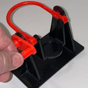 May include: A black plastic toy with a red rubber band and a red plastic handle. The toy is designed to launch a small object.