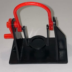 May include: A black plastic toy with a red plastic loop. The toy has a circular opening in the centre and two arms that move up and down.