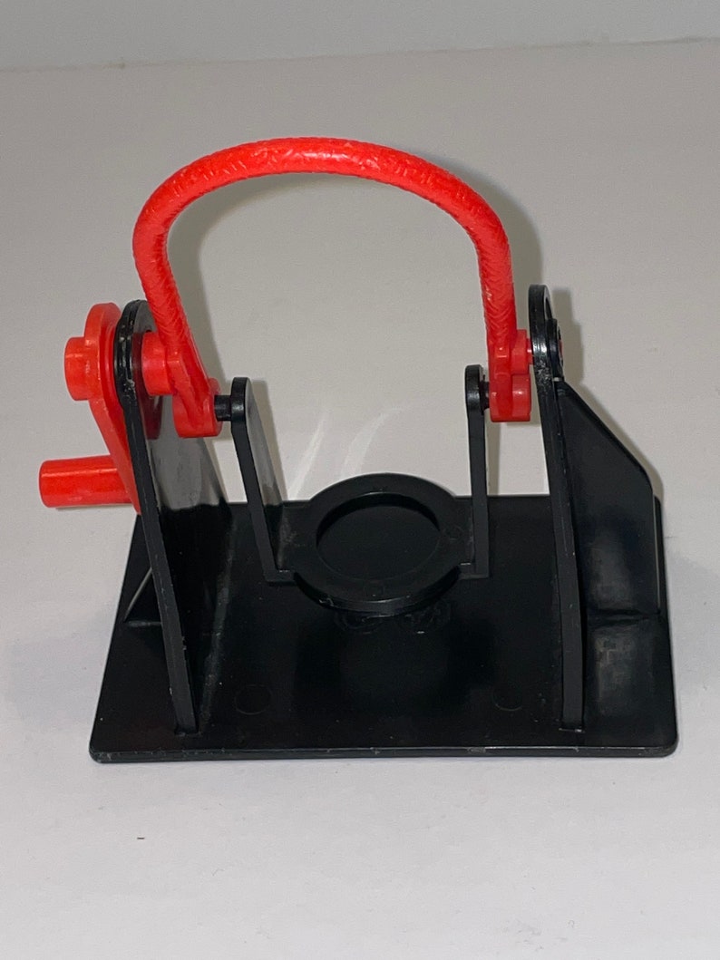 May include: A black plastic toy with a red plastic loop. The toy has a circular opening in the centre and two arms that move up and down.