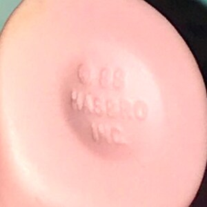 Puede incluir: Primer plano de un juguete de pl&aacute;stico rosa con el texto "1988 Hasbro Inc." impreso en la parte inferior.
