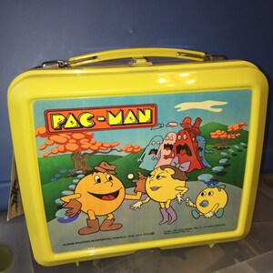 May include: Yellow plastic lunchbox featuring Pac-Man characters. The lunchbox has a black handle and a blue and green scene with Pac-Man, Ms. Pac-Man, and ghosts. The text "PAC-MAN" is printed in red on the front of the lunchbox.