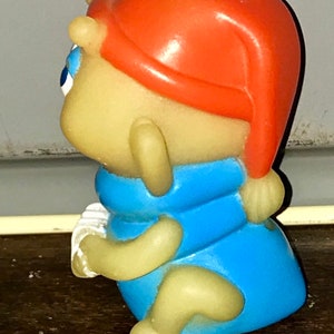 Puede incluir: Una figura de juguete de pl&aacute;stico amarillo y azul de una criatura que lleva un sombrero rojo. La figura sostiene un objeto blanco en su mano.