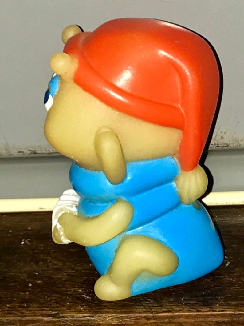 Puede incluir: Una figura de juguete de pl&aacute;stico amarillo y azul de una criatura que lleva un sombrero rojo. La figura sostiene un objeto blanco en su mano.