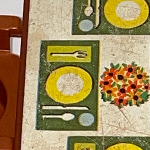 May include: A brown wooden table with a white and green patterned tabletop. The tabletop has a design of plates, forks, knives, spoons, and a floral centerpiece.