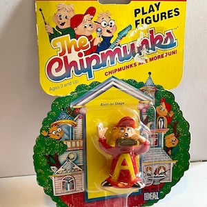 May include: A plastic Alvin the Chipmunk figure playing a harmonica. The figure is red and yellow with a large letter "A" on the front. The figure is packaged on a yellow and red card with the words "The Chipmunks" and "Chipmunks Are More Fun!" The card also includes a picture of the Chipmunks playing musical instruments.