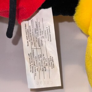 May include: Close-up of a white tag with black text attached to a red, black, and yellow plush toy. The tag states "Not to be removed until delivered to the consumer. This label is affixed in compliance with Provincial law. This article contains NEW MATERIAL ONLY. Made by Ont. Reg. No. 99Z 7475. CONTENTS Polyester fiber & Plastic stiffener."