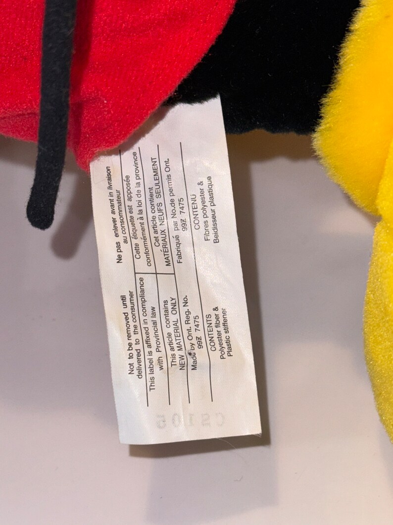 May include: Close-up of a white tag with black text attached to a red, black, and yellow plush toy. The tag states "Not to be removed until delivered to the consumer. This label is affixed in compliance with Provincial law. This article contains NEW MATERIAL ONLY. Made by Ont. Reg. No. 99Z 7475. CONTENTS Polyester fiber & Plastic stiffener."