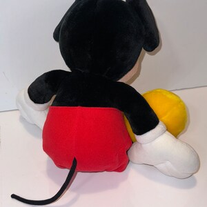 May include: A plush black and red Mickey Mouse toy sitting with a yellow plush toy. The Mickey Mouse toy has a black tail and white gloves.