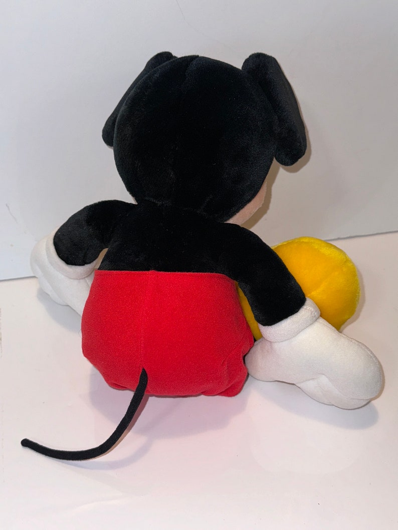 May include: A plush black and red Mickey Mouse toy sitting with a yellow plush toy. The Mickey Mouse toy has a black tail and white gloves.