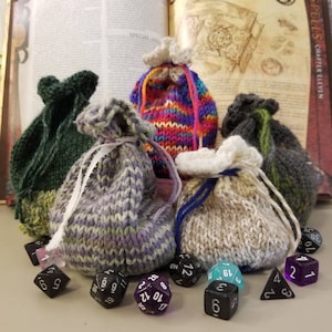 May include: Five knitted bags in various colors, including green, purple, pink, and beige, with a white drawstring. The bags are filled with dice, including black, purple, and blue dice. The dice are numbered 1 through 20.