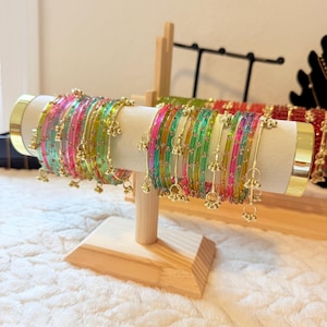 May include: A set of translucent bangles in vibrant shades of pink, green, and yellow, featuring small metallic accents and dangling charms. These circular bracelets are arranged on a light-coloured cylindrical display stand with gold-toned ends.