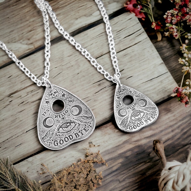 Two silver necklaces with pendants shaped like a planchettes from Ouija boards. Each pendant features a crescent moon, an eye, and the words "Yes", "No", "Hello", "Goodbye". one pendant is larger than the other.
