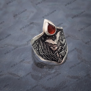 Könnte beinhalten: Silberner Ring mit schildförmigem Design, das ein Schloss, Fledermäuse und eine Berglandschaft zeigt. Er hat einen roten, tropfenförmigen Edelstein. Der Ring ist mit den Worten "WYRd Charm" graviert.
