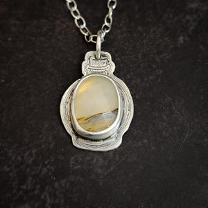 May include: A silver pendant necklace featuring an oval-shaped, light-colored stone with brown and gray inclusions. The stone is set in a decorative silver frame, suspended from a silver chain. The pendant has an antique, vintage aesthetic.