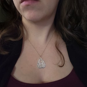 A silver necklace with a pendant shaped like a planchette from a Ouija board. The pendant has the words "hello", "goodbye", "yes", and "no" inscribed on it, along with other symbols.