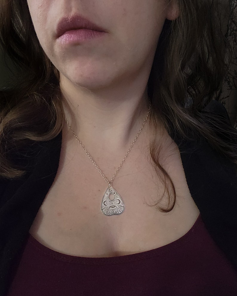 A silver necklace with a pendant shaped like a planchette from a Ouija board. The pendant has the words "hello", "goodbye", "yes", and "no" inscribed on it, along with other symbols.