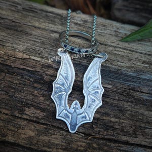 Könnte beinhalten: Eine silberne Fledermaus-Anhänger-Halskette mit passendem Ring. Der Anhänger zeigt ein detailliertes Fledermaus-Design mit ausgebreiteten Flügeln. Der Ring ist silberfarben mit kleinen blauen Steinen. Der Text "wyrharm" ist sichtbar.