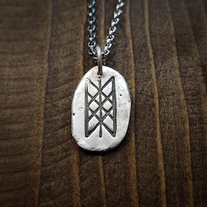 Peut inclure: Un collier avec un pendentif argenté de forme ovale et texturé. Le pendentif présente un motif géométrique de lignes verticales et diagonales. La chaîne est d'un argent plus foncé. Le fond est une surface en bois sombre.