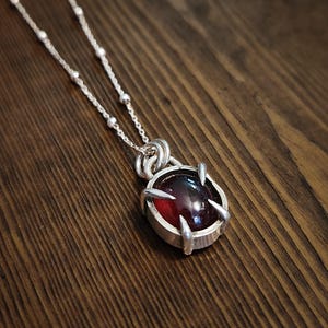 Könnte beinhalten: Eine silberne Anhängerkette mit einem tiefroten, ovalen Cabochon-Edelstein, der von vier klauenartigen Krappen gehalten wird. Der Anhänger hängt an einer feinen Silberkette mit kleinen, gleichmäßig verteilten runden Perlen.