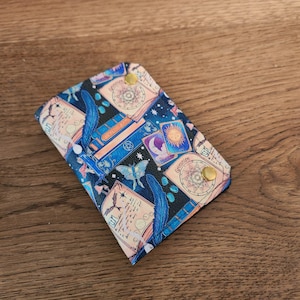 May include: A rectangular wallet featuring a dark blue background with a pattern of celestial motifs, including a sun, a moth, feathers, and cards labeled Grimoire. The wallet is secured with two gold-colored metal snap buttons on the front flap.