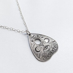 Silver necklace with a silver pendant shaped like a Ouija board planchette. The pendant has the words "YES", "NO", "HELLO", and "GOODBYE" inscribed around the edge. The center of the pendant features a circle with an eye inside.