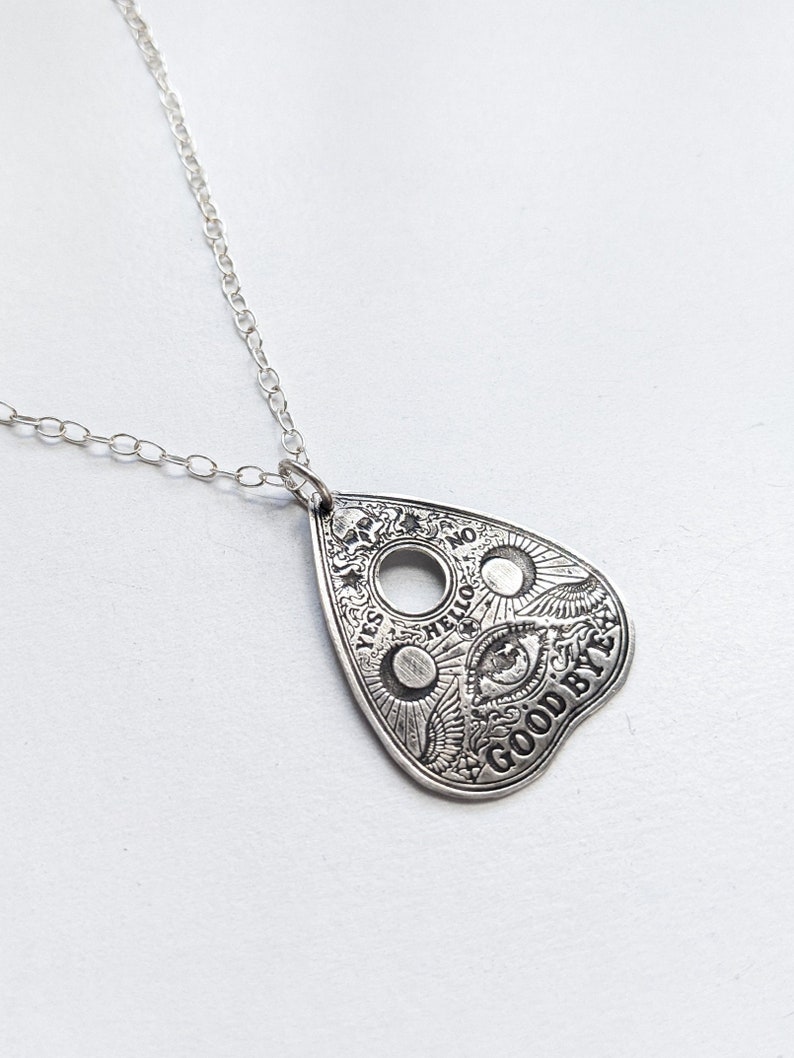Silver necklace with a silver pendant shaped like a Ouija board planchette. The pendant has the words "YES", "NO", "HELLO", and "GOODBYE" inscribed around the edge. The center of the pendant features a circle with an eye inside.