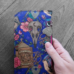 Può includere: Un portafoglio blu con un motivo floreale e a tema occulto. Il design include un teschio, forbici, un libro intitolato "Book of Shadows" e una tavola Ouija. Il portafoglio ha un bordo rosa e un ciondolo argentato.