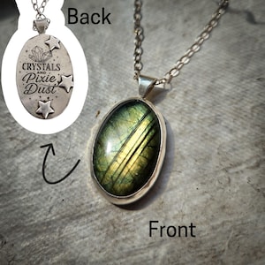Puede incluir: Collar de plata con un colgante ovalado. La parte delantera del colgante presenta una piedra verde y dorada. La parte trasera del colgante tiene las palabras "Crystals and Pixie Dust" con diseños de estrellas y cristales.