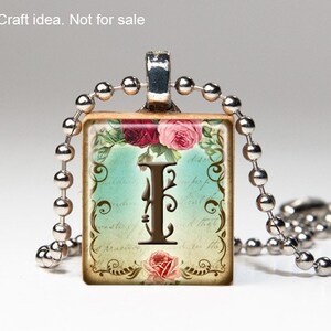 May include: A silver chain necklace with a square pendant featuring a vintage-style illustration of the letter I surrounded by floral designs and a blue background. The pendant has a brown border and the text "I am the" is visible on the bottom.