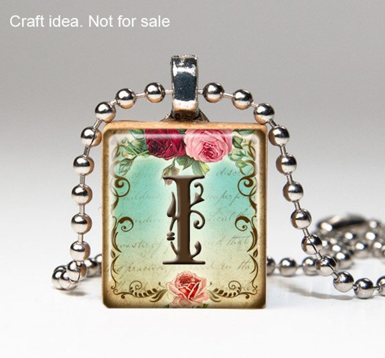 May include: A silver chain necklace with a square pendant featuring a vintage-style illustration of the letter I surrounded by floral designs and a blue background. The pendant has a brown border and the text "I am the" is visible on the bottom.
