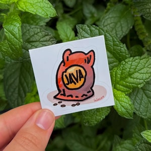 May include: A square sticker featuring a cartoon coffee bean bag design. The bag is red-orange with a yellow oval that says "JAVA" in black. Several coffee beans are scattered below the bag. The sticker is held against a background of green leaves.