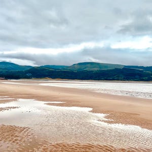 Puede incluir: Una amplia playa de arena con una gran masa de agua en el fondo. El agua está tranquila y refleja el cielo nublado. En la distancia, una cadena de colinas verdes se eleva en el horizonte.