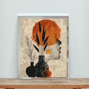 Puede incluir: Impresión artística abstracta con el perfil de una mujer y un sol naranja. Plantas negras silueteadas en jarrones en primer plano. La obra está enmarcada en un marco plateado y descansa sobre una superficie de madera.