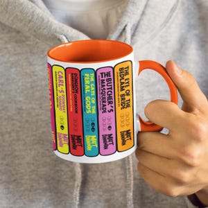 May include: A white ceramic mug with an orange interior and handle, featuring a graphic print of colourful book spines titled The Eye of the Bedlam Bride, The Butcher's Masquerade, The Gate of the Feral Gods, Dungeon Crawler Carl's Cookbook, and Carl's Doomsday Scenario.