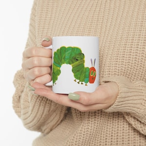 May include: White ceramic mug featuring a colorful illustration of a green caterpillar with a red head and purple antennae. The caterpillar design is a whimsical, cartoon-style image. The mug has a white handle.