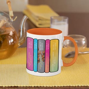 Puede incluir: Una taza de cerámica blanca con interior y asa de color naranja, que presenta un gráfico de cinco lomos de libros coloridos. El texto en los lomos dice: A Court of Wings and Ruin, A Court of Frost and Starlight, A Court of Silver Flames, A Court of Splintered Harmony y A Court of Forgotten Melody, todos de Sarah J. Maas.
