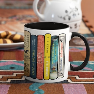 Könnte beinhalten: Ein weißer Keramikbecher mit schwarzem Innenraum und Henkel, bedruckt mit einer Grafik von fünf Buchrücken. Die Titel umfassen Fourth Wing, Throne of Glass, Vatto, Yellowface, Under the Whispering Door und Demon Copperhead mit den Autorennamen.