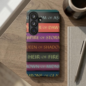 Könnte beinhalten: Smartphone-Hülle mit einem Stapel bunter Buchrücken. Titel wie "Throne of Glass" und "Heir of Fire" sind zu sehen. Die Hülle liegt auf einem Holztisch, neben einem Notizbuch, Stift und einer Kaffeetasse.