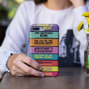 May include: A phone case featuring a stacked book spine design with titles from the Dungeon Crawler Carl series, including A Parade of Gambles, This Inevitable Ruin, The Eye of the Bedlam Bride, The Butcher's Masquerade, The Gate of the Feral Gods, and Carl.