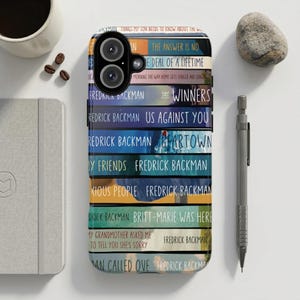 May include: A smartphone case featuring a stack of book spines with titles by Fredrick Backman. The case has a black camera module and a colorful design with titles like "Winners" and "Us Against You".