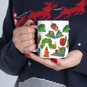 May include: White ceramic mug featuring colorful illustrations of a caterpillar, fruits, and butterflies. The design includes a green caterpillar, red apple, and a watermelon slice. The mug is held by a person wearing a navy blue sweater.